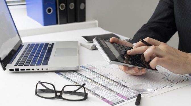 Pessoa fazendo contas de clientes inadimplentes em uma calculadora, em frente a um notebook e sobre papéis.