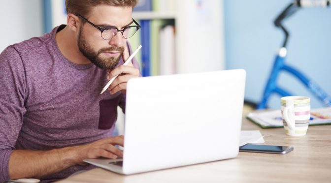 Homem ruivo e com barba sentado em frente a um notebook, pesquisando nomes para provedor de internet.