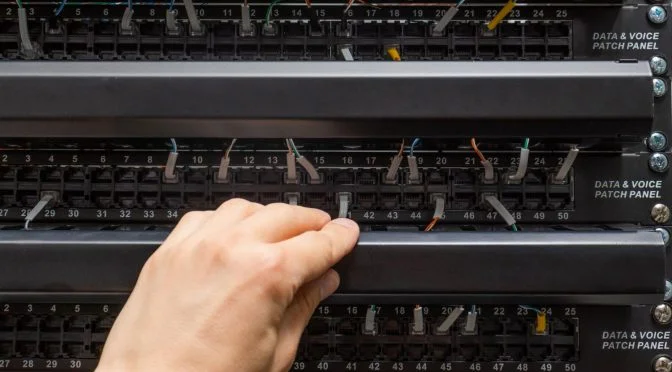 Patch panel preto com cabos conectados e uma mão masculina posicionada no meio da estrutura.