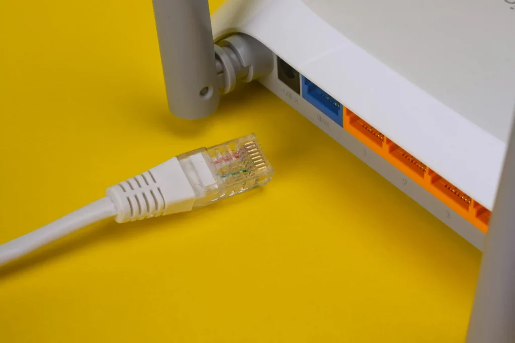 Roteador branco semelhante a um aparelho Simet Box, com cabo ao lado, em um fundo amarelo.